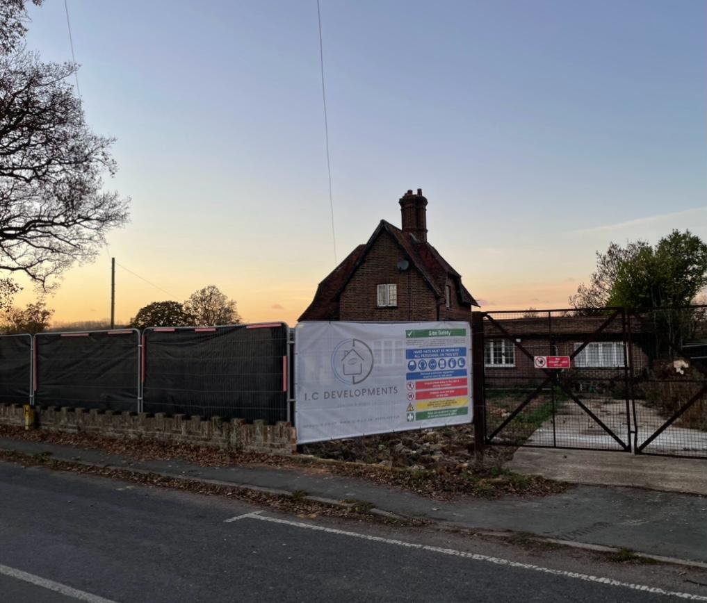 A hoarded I.C Developments site at dusk in front of an older brick and clay tile house in Surrey