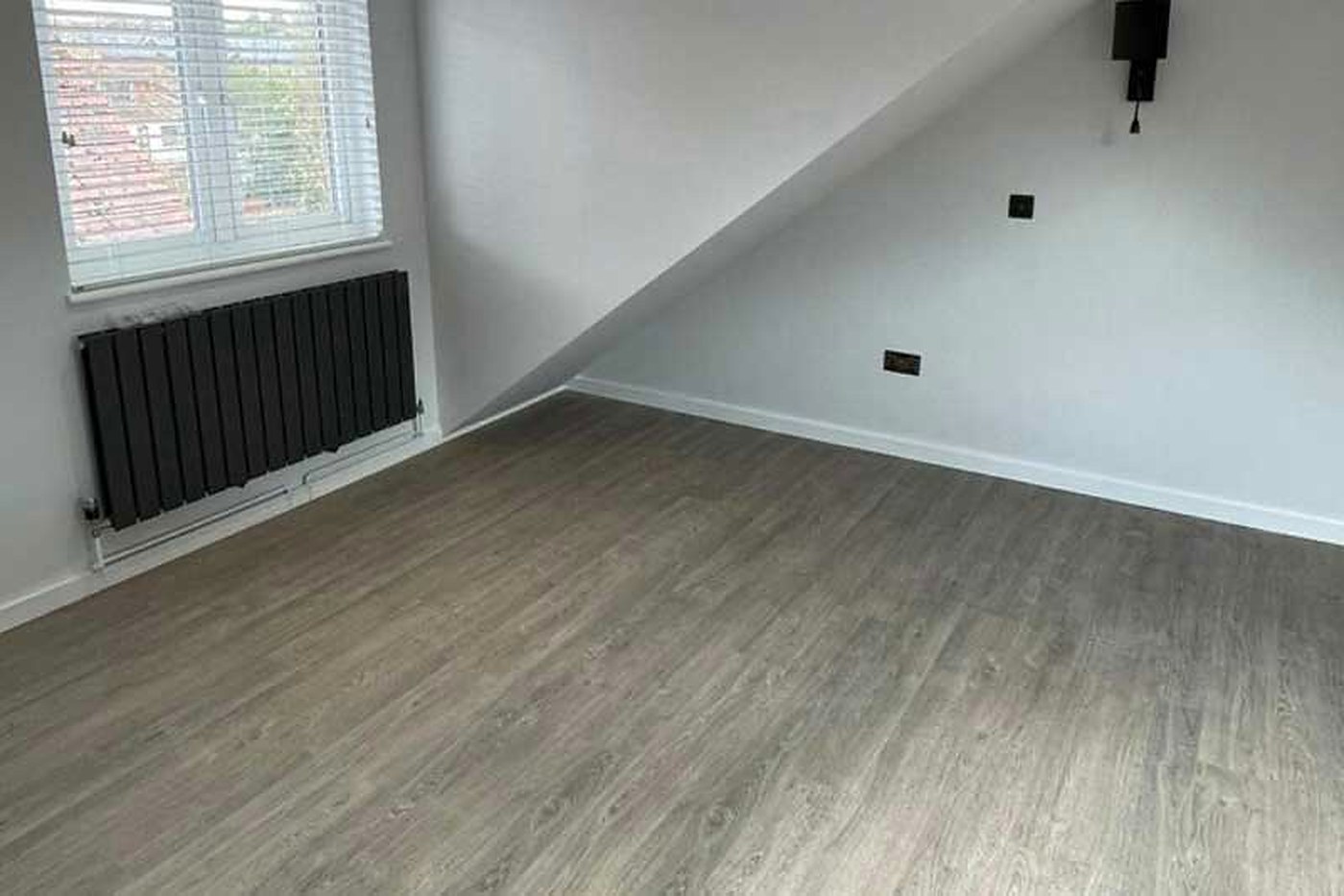 A finished loft room with a sloping ceiling, a new black radiator under the window and timber effect flooring